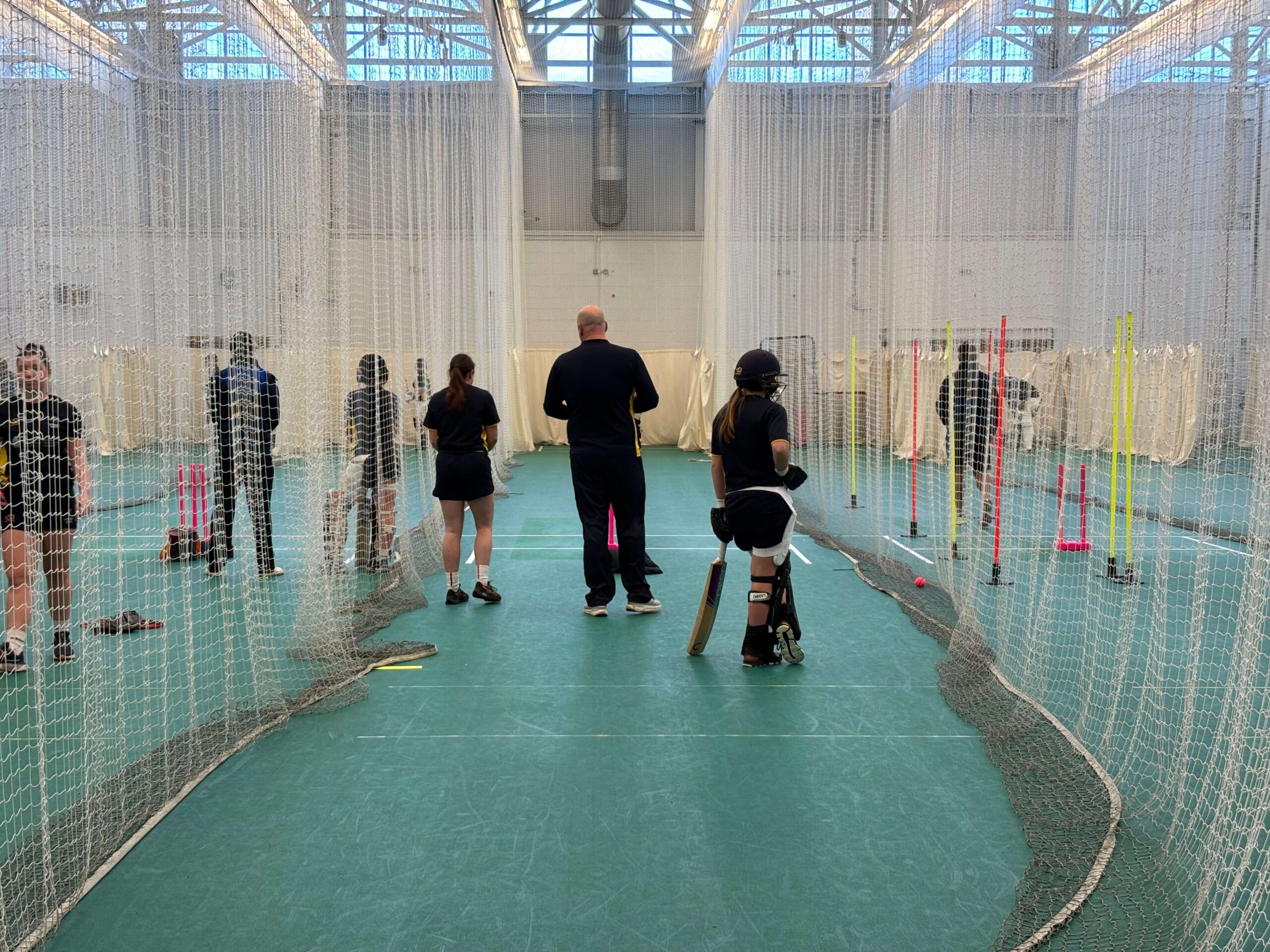 Young women in Glamorgan County Cricket Club kit turned away from camera. In sports hall surrounded by nets