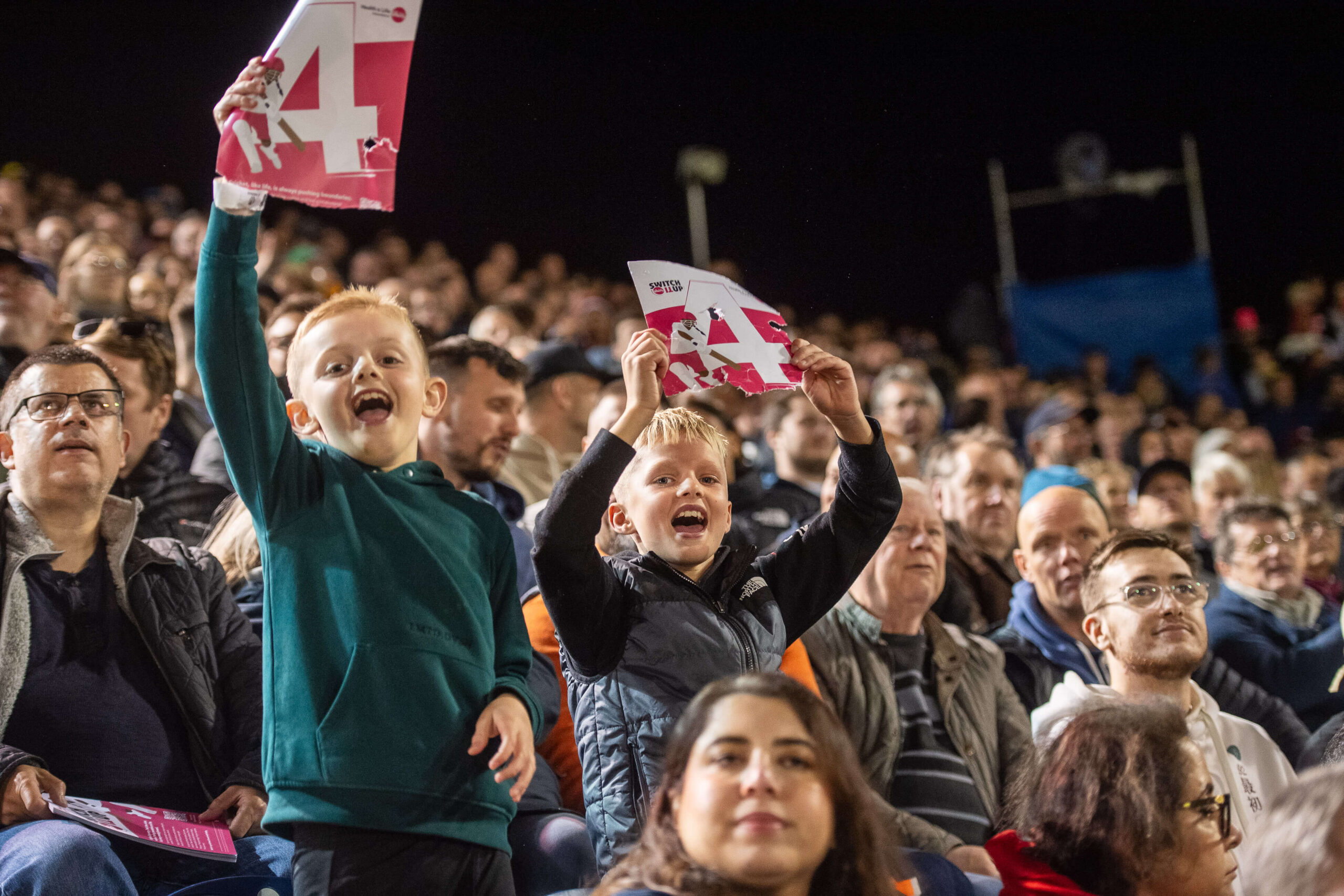 Crowd of cricket fans watching a match. Two children on shoulders look into camera waving signs